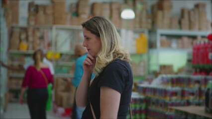 Female consumer looking for products on shelf at grocery store, considering merchandise for sale,...