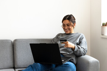 young black-haired woman shopping online from the sofa at home with the computer and credit or debit card