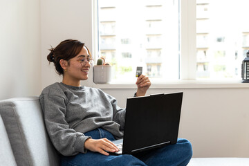 young black-haired woman shopping online from the sofa at home with the computer and credit or debit card