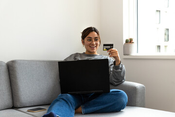 young black-haired woman shopping online from the sofa at home with the computer and credit or debit card