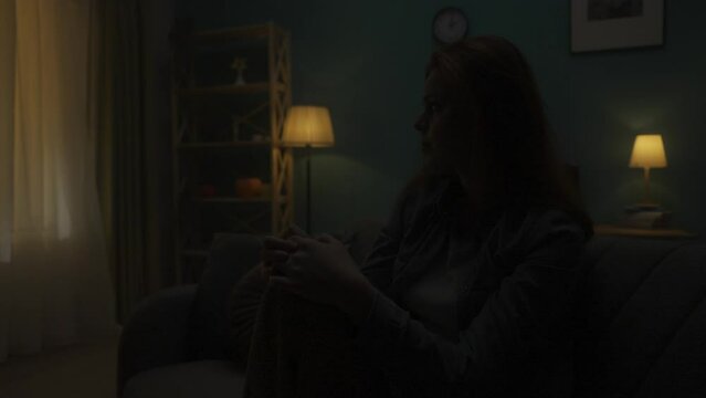 Devastated woman sitting on a sofa in a dark room close up. A woman desperately thinks about personal problems, is going through a crisis. Change of day and night. HDR BT2020 HLG Material.