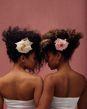 Two females with flowers in their heads standing head to head with closed eyes against a pink wall