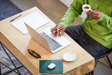 Cropped top view image of african man in green sweater sitting at table, drinking coffee, working on laptop online. Distance learning and work. Concept of business and education, freelance job