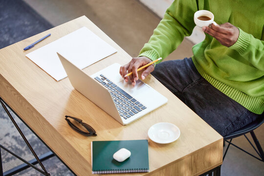 Cropped top view image of african man in green sweater sitting at table, drinking coffee, working on laptop online. Distance learning and work. Concept of business and education, freelance job