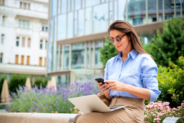 Brunette haired woman sitting on a bench in the city and using a laptop and smartphone