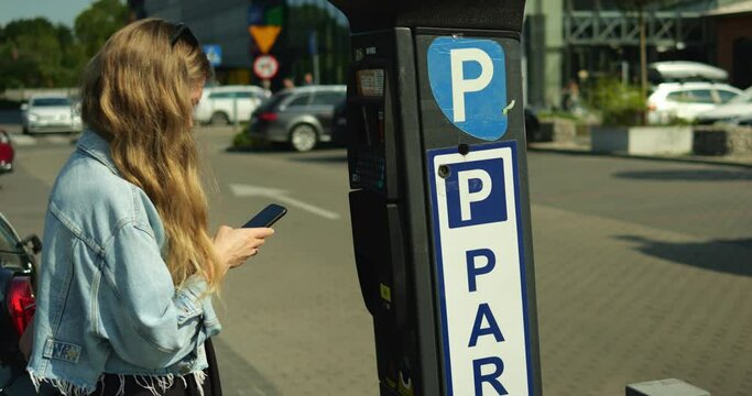 Young beautiful woman in denim jacket stand at parking meter with smartphone. Female student with long brown hair prepares to pay contactless with mobile phone at outdoors car parking lot on sunny day