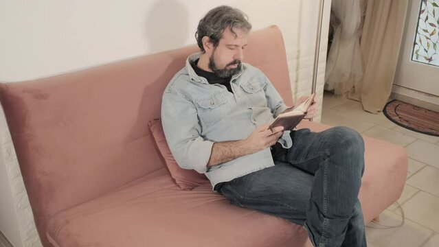 A middle-aged man reading an old book on the sofa at home, slowly falling asleep, but waking up just in time.

