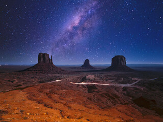 Obraz na płótnie Canvas View of the 3 monoliths of Monument Valley at night, with starry sky and milky way, and a car passing on the road below