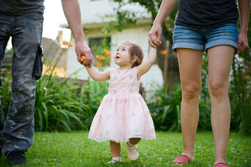 Fototapeta premium Lovely family walking holding hands, adopted child being supported by loving parents. Girl holds hands of her parents and jumps. Cropportrait of the cute adorable small girl in dress.
