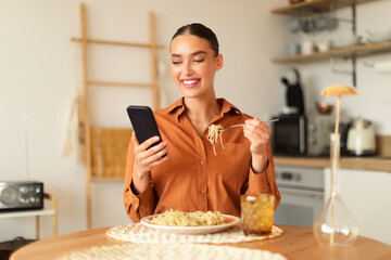 Gadget addiction. Happy young caucasian woman having lunch in kitchen at home, eating pasta and...