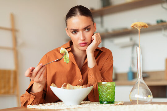 Thoughtful young woman eating caesar salad and having lack of appetite, sitting at table in kitchen interior, copy space