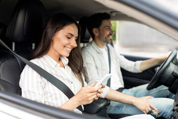 Happy millennial couple sitting inside white car, woman holding smartphone