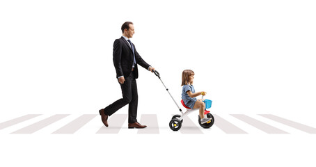 Full length profile shot of a father pushing a girl on a tricycle at a pedestrian crossing