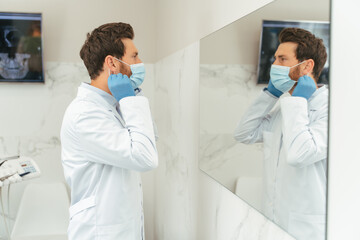Fototapeta premium Portrait of handsome middle aged dentist wearing white coat latex gloves and wearing protective mask