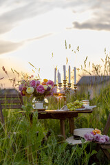 Romantic surprise, marriage proposal. Elegant decoration. Private wedding party for two, table set with floral decor, chandelier, fruits, wine in field or meadow. Summer, sunset, golden hour.
