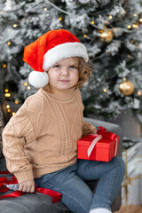 Fototapeta na wymiar Portrait of a cute little girl in a beautiful knitted sweater ad a red Santa hat near decorated Christmas and new year tree at home. Smiling, cheerful and happy, holding red gift box