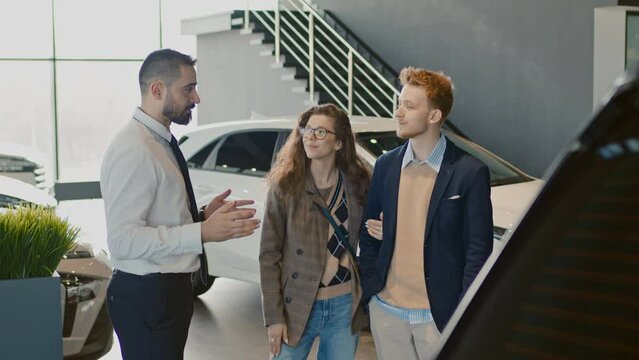 Medium arc shot of biracial salesman talking to young Caucasian husband and wife in showroom, explaining car characteristics and features and recommending vehicle modelsMedium arc shot of biracial sal