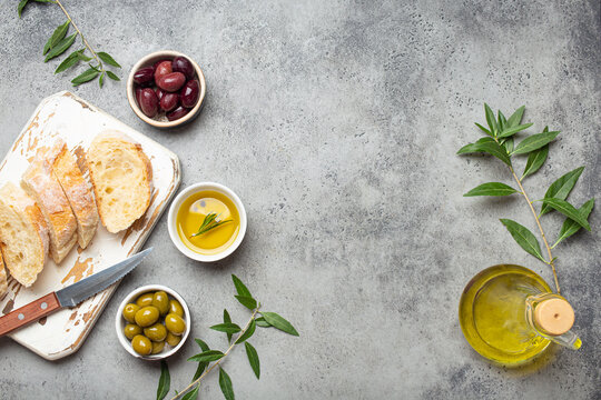 Sliced Fresh Ciabatta On Cutting Kitchen Board, Green And Brown Olives, Olive Oil With Rosemary, Olive Tree Branches On Gray Concrete Stone Rustic Background From Above Copy Space