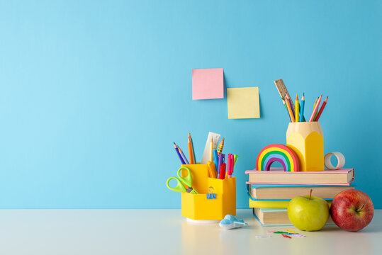 Classroom essentials on display: side view photo of desk adorned with school supplies, pencil holders, colorful pens, rulers, books, sticky notes, plasticine, and apples against a blue wall backdrop