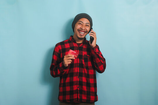 Excited Young Asian Man With A Beanie Hat And A Red Plaid Flannel Shirt Is Smiling While Holding A Credit Card And Talking On His Smartphone To Make An Online Payment, Isolated On A Blue Background