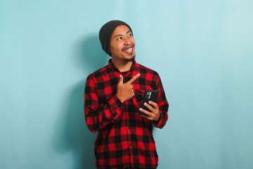 Young Asian man with a beanie hat and a red plaid flannel shirt is pointing his finger right at copy space while holding a mobile phone, isolated on a blue background