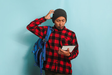 A surprised young Asian student with a beanie hat and a red plaid flannel shirt, wearing a backpack, scratching his head appears unhappy and confused, isolated on a blue background