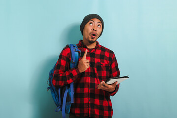 An Excited smiling young Asian student with a beanie hat and a red plaid flannel shirt, wearing a backpack, is pointing finger up at copy space while standing against a blue background