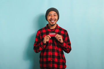 Excited young Asian man with a beanie hat and red plaid flannel shirt is holding bank credit cards in his hands while standing against a blue background. Finance, loan, savings concept