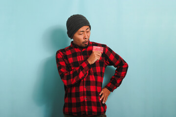 Young Asian man with a beanie hat and red plaid flannel shirt is holding bank credit cards in his hands, with a surprised expression on his face while standing against a blue background.