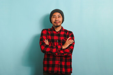 A confident young Asian man with a beanie hat and a red plaid flannel shirt is looking at the camera with his arms crossed. He is isolated on a blue background