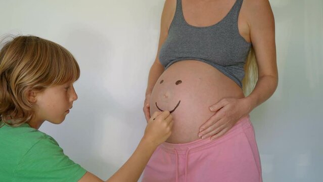 A little boy drawing a smiling face on his pregnant mother's belly. The scene exudes warmth and tenderness, showcasing the special bond between the mother and her child. The child's innocent and