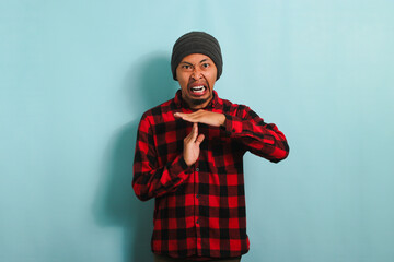 Angry Young Asian man with beanie hat and red plaid flannel shirt demonstrating a time out or enough gesture with his arms, isolated on a blue background