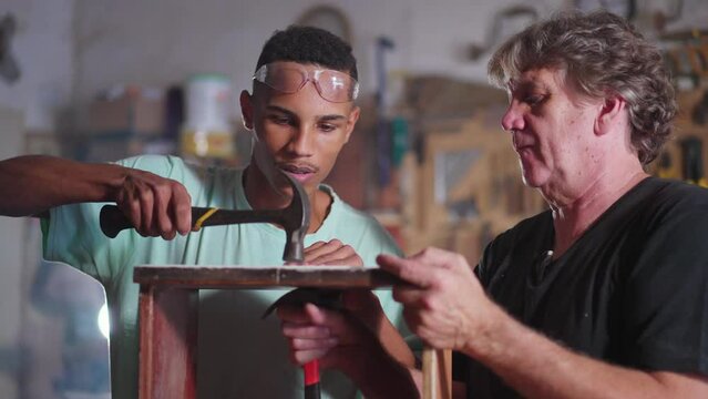 Master carpenter orienting young apprentice to do woodwork at carpentry workshop. Senior man teaching young student to work at business