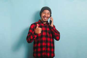 Excited Young Asian man with a beanie hat and a red plaid flannel shirt is showing a thumbs up while talking on his mobile phone, isolated on a blue background
