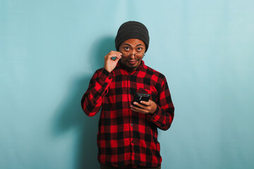 Young Asian man with a beanie hat and a red plaid flannel shirt is staring through his eyeglasses while holding a phone, isolated on a blue background - obrazy, fototapety, plakaty