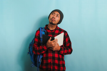 A pensive young Asian student with a beanie hat and a red plaid flannel shirt, wearing a backpack, is deep in thought, looking up at the copy space while standing against a blue background