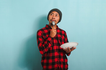 Pensive Young Asian man with beanie hat and red plaid flannel shirt holding a spoon and an empty white plate in his hand, thinking about what to eat, isolated on a blue background