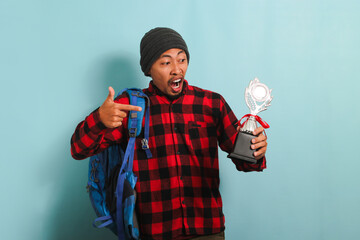 Excited Young Asian man student wearing a backpack, beanie hat, and red plaid flannel shirt, proudly pointing a victory trophy in his hand, isolated on a blue background