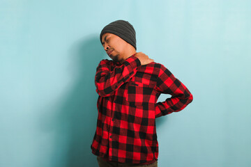 A young Asian man with a beanie hat and a red plaid flannel shirt massages his neck or shoulders,...