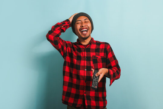 Happy young Asian man with a beanie hat and red plaid flannel shirt bursting out laughing while watching TV, isolated on a blue background