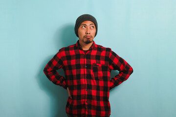 A bored young Asian man wearing a beanie hat and a red plaid flannel shirt stands with an arm...