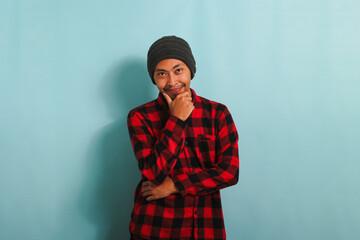 A confident young Asian man with a beanie hat and a red plaid flannel shirt is holding his hand on his chin and smiling. He is isolated on a blue background