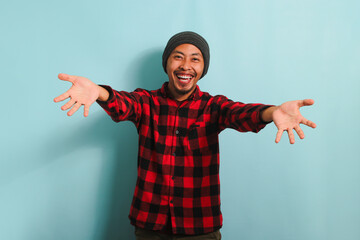 Smiling young Asian man wearing a beanie hat and a red plaid flannel shirt is beckoning with both hands, offering a hug with a come here gesture, inviting, welcoming, isolated on blue background