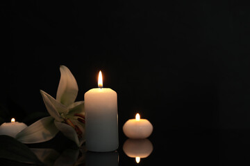 Fototapeta na wymiar White lily and burning candles on black mirror surface in darkness, space for text. Funeral symbols