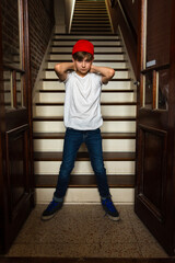 Boy in red cap and blue jeans standing in the doorway to a stairwell with arms behind his head