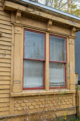 The exterior wall of a yellow wooden house with a large double hung window. The glass window has a curtain and blind. The pattern on the outside of the wall is small scallop shapes and horizontal wood
