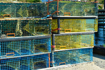 Multiple yellow lobster pots or traps stacked high along a wharf. Containers are made from yellow wire and nylon netting and in a square shape. There's no bait in the pots, all are empty.