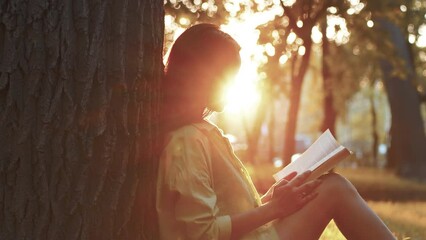 Young woman sitting under a tree in the city park in summer and reading a book in the backlight of sunset. Concept of rest and appeasement in bustle urban life - Powered by Adobe