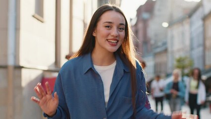 Hello. Young pretty woman smiling friendly at camera, waving hands gesturing hi, greeting or goodbye, welcoming with hospitable expression outdoors. Happy lovely girl walking in urban city town street
