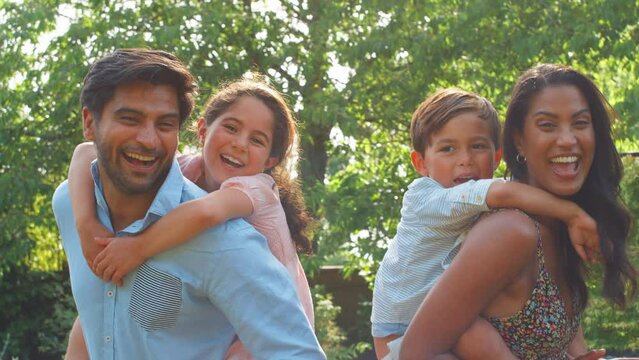 Portrait of family of four having fun outdoors in countryside or garden with parents giving children piggyback rides - shot in slow motion